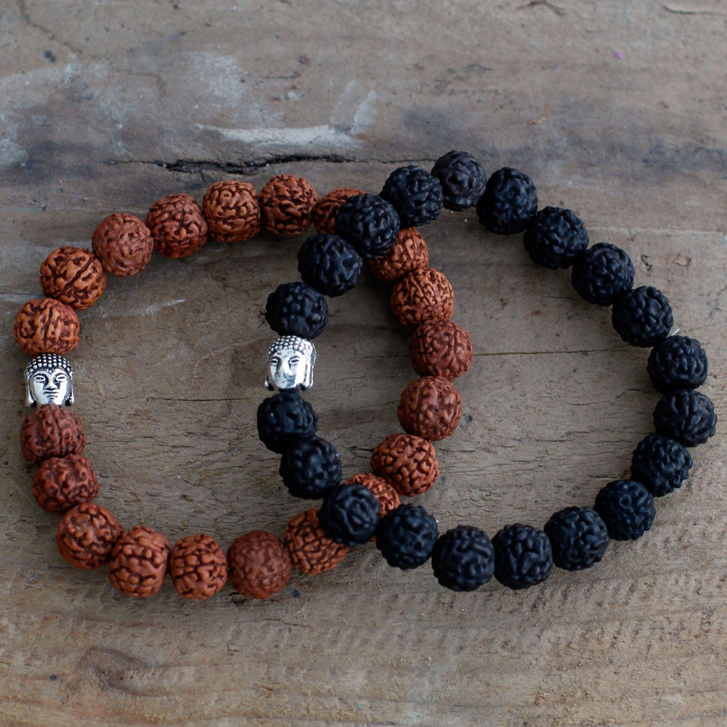 Deux bracelets mala en perles de rudraksha noir et brun avec tête de Bouddha posés sur fond bois naturel
