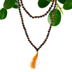 Collier mala 108 perles en pierre naturelle oeil de tigre avec pompon orange, suspendu avec feuilles vertes en arriere plan, bijou spirituel pour meditation chakra et bien etre