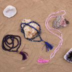 Trois malas en pierres naturelles poses sur fond sable avec cristaux decoratifs, incluant quartz rose, amethyste et lapis lazuli avec pompons colores pour meditation et bien etre