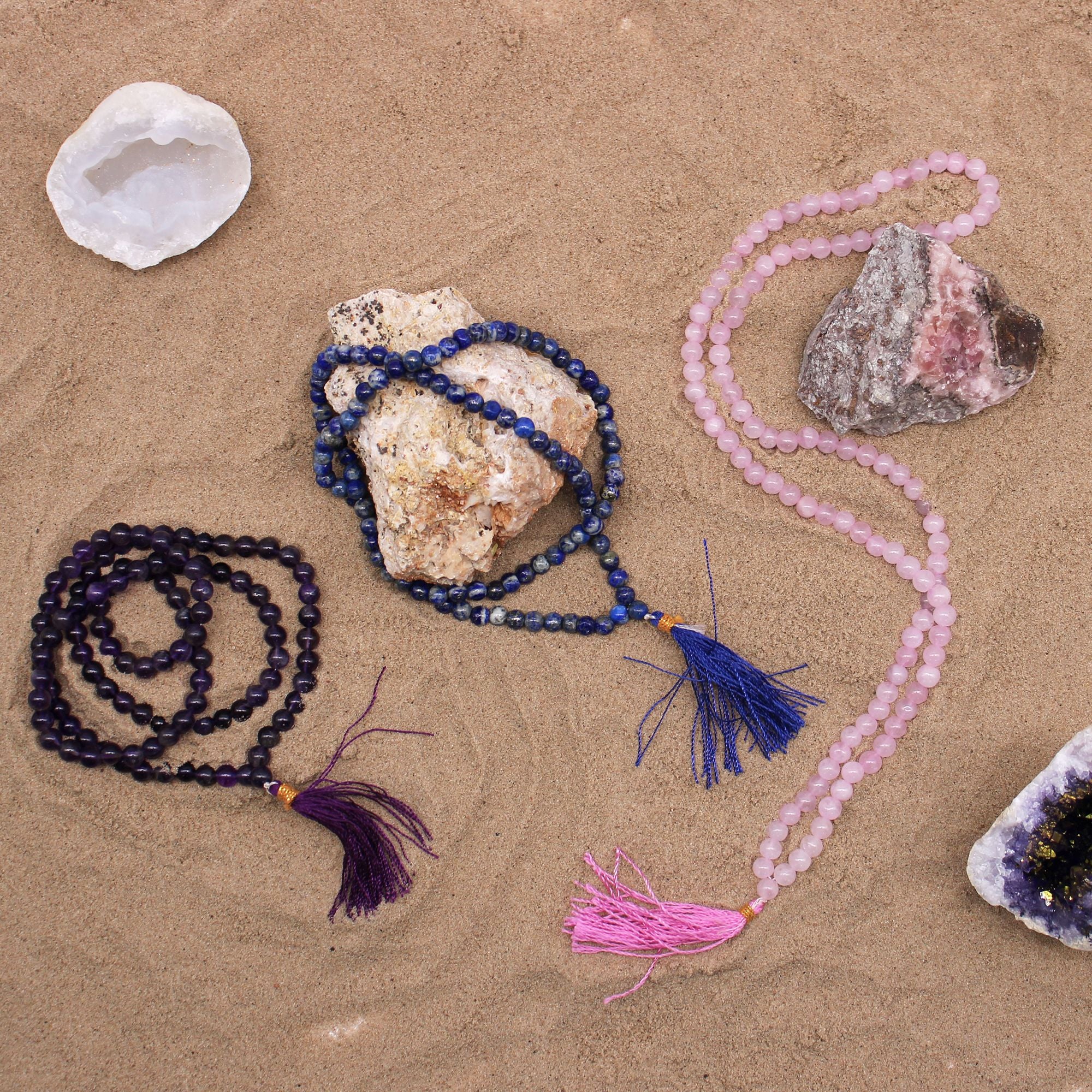 Trois malas en pierres naturelles poses sur fond sable avec cristaux decoratifs, incluant quartz rose, amethyste et lapis lazuli avec pompons colores pour meditation et bien etre