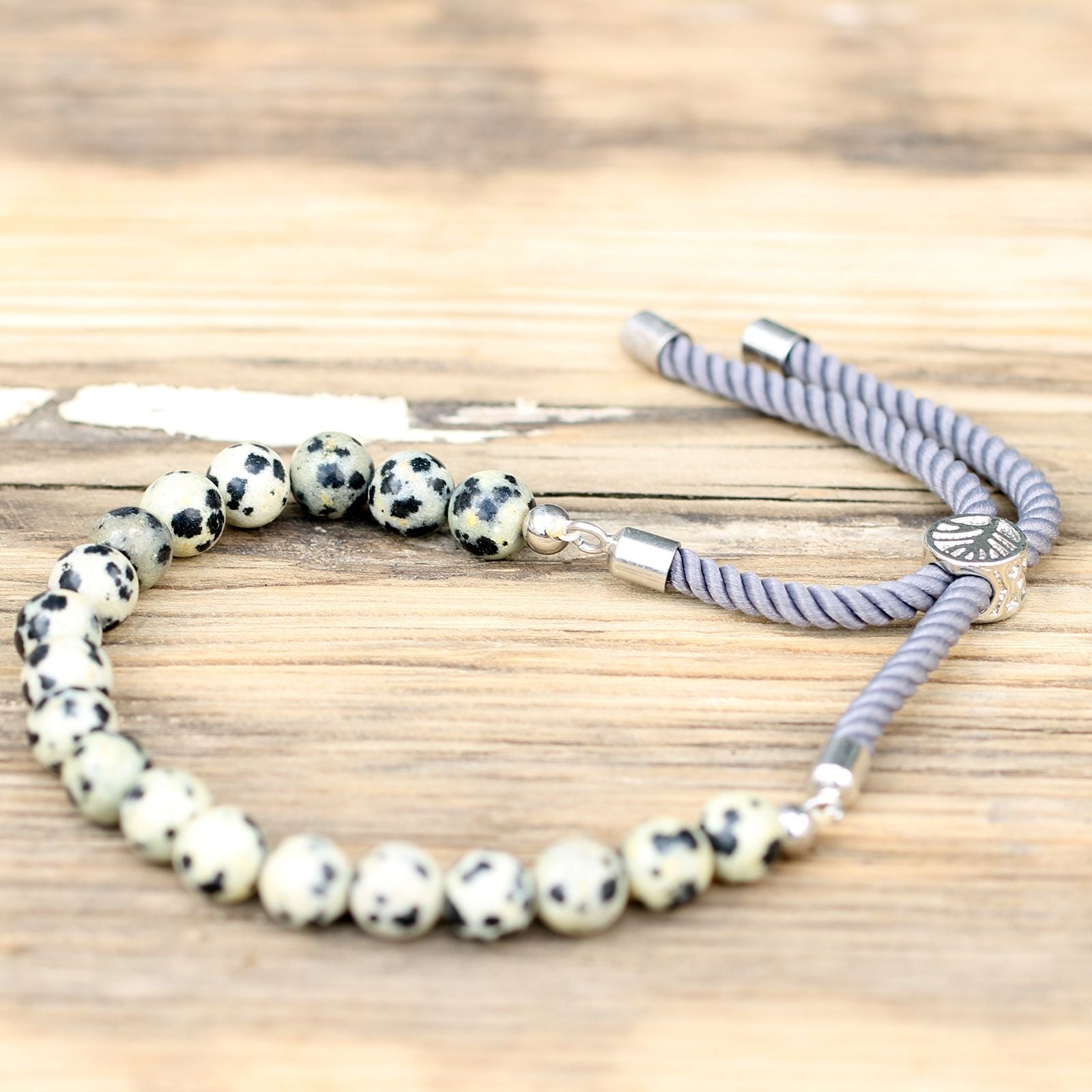 Bracelet en pierre naturelle jaspe dalmatien ajustable avec cordon gris et perles noir et blanche posé sur table en bois